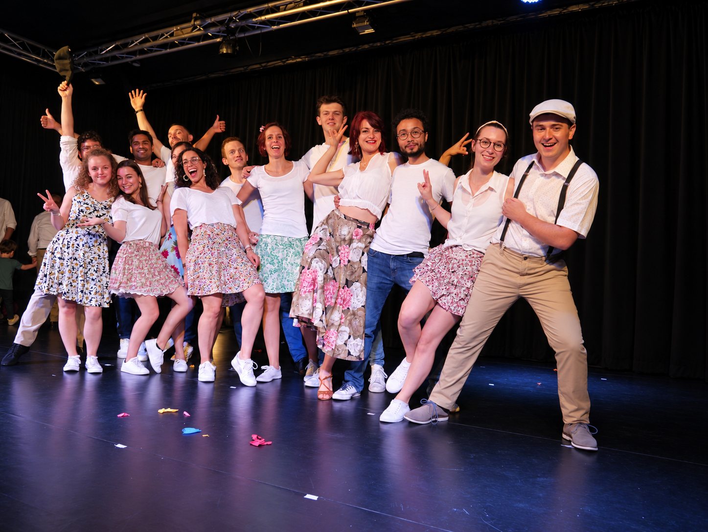 Un groupe de danseurs souriants, vêtus de blanc et de jupes à fleurs, pose sur une scène sombre parsemée de confettis.