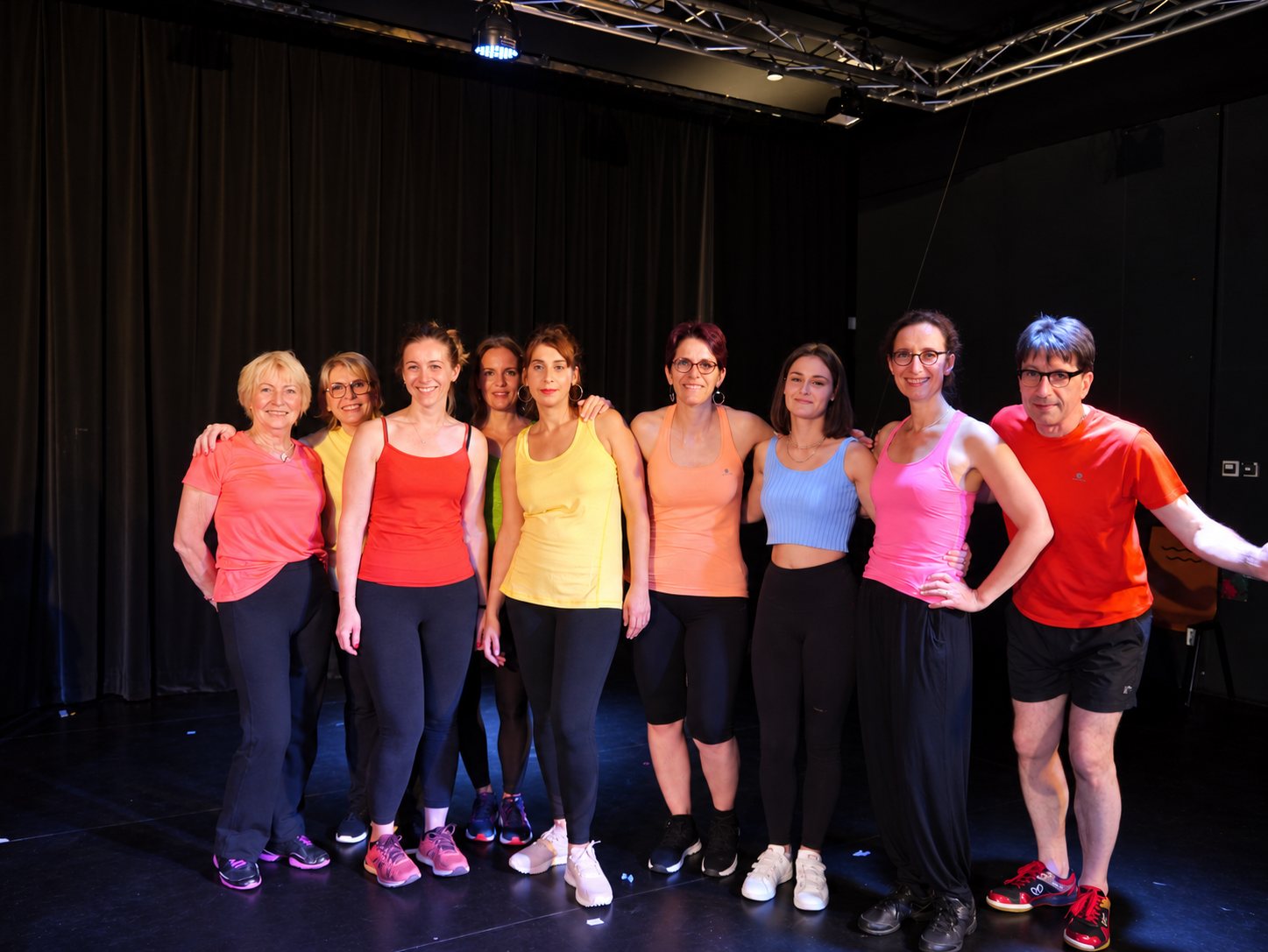 Neuf personnes en vêtements de sport orange, rose et jaune posent côte à côte sur une scène de théâtre sombre, devant un rideau noir.