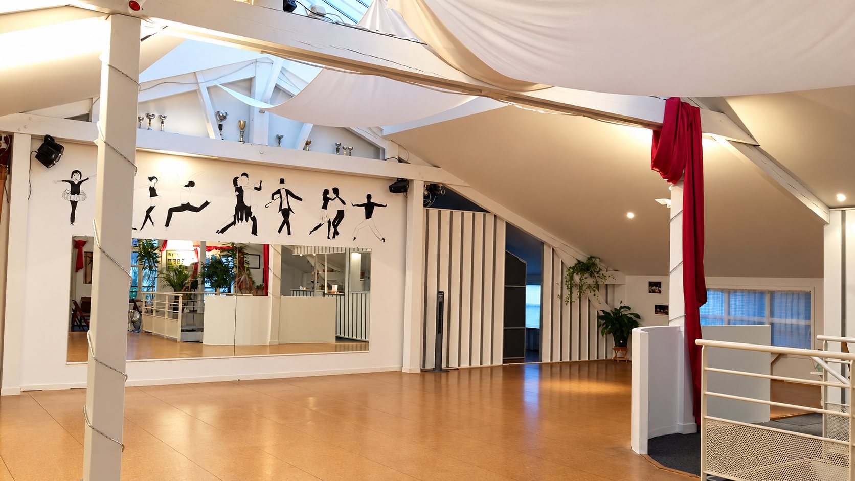 Intérieur d’une salle de danse au parquet clair, avec un grand miroir, des silhouettes de danseurs peintes sur le mur et un rideau rouge.