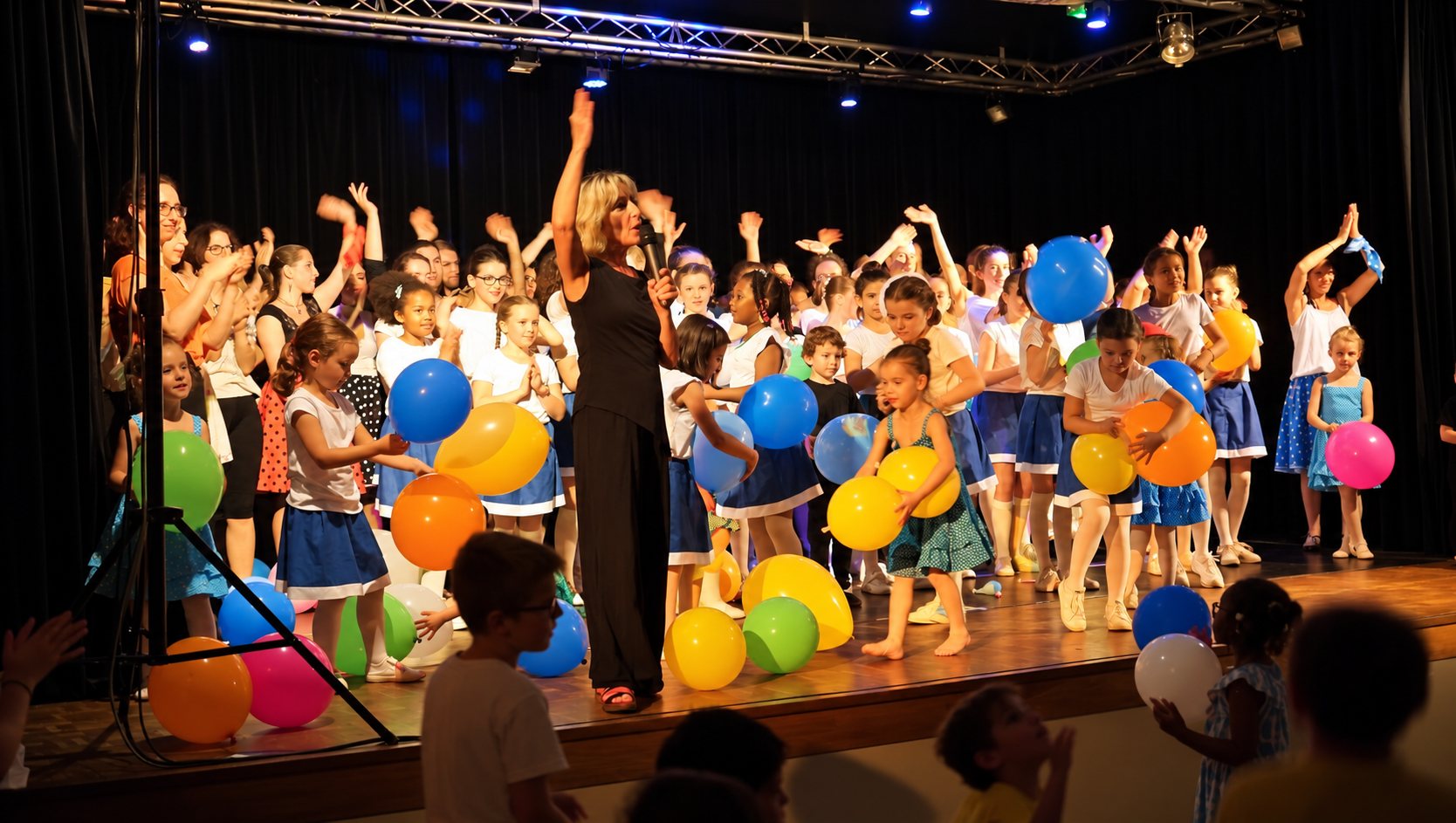 Une femme au micro se tient au centre d’une scène entourée d’enfants qui lèvent les bras et tiennent des ballons colorés pendant un spectacle.